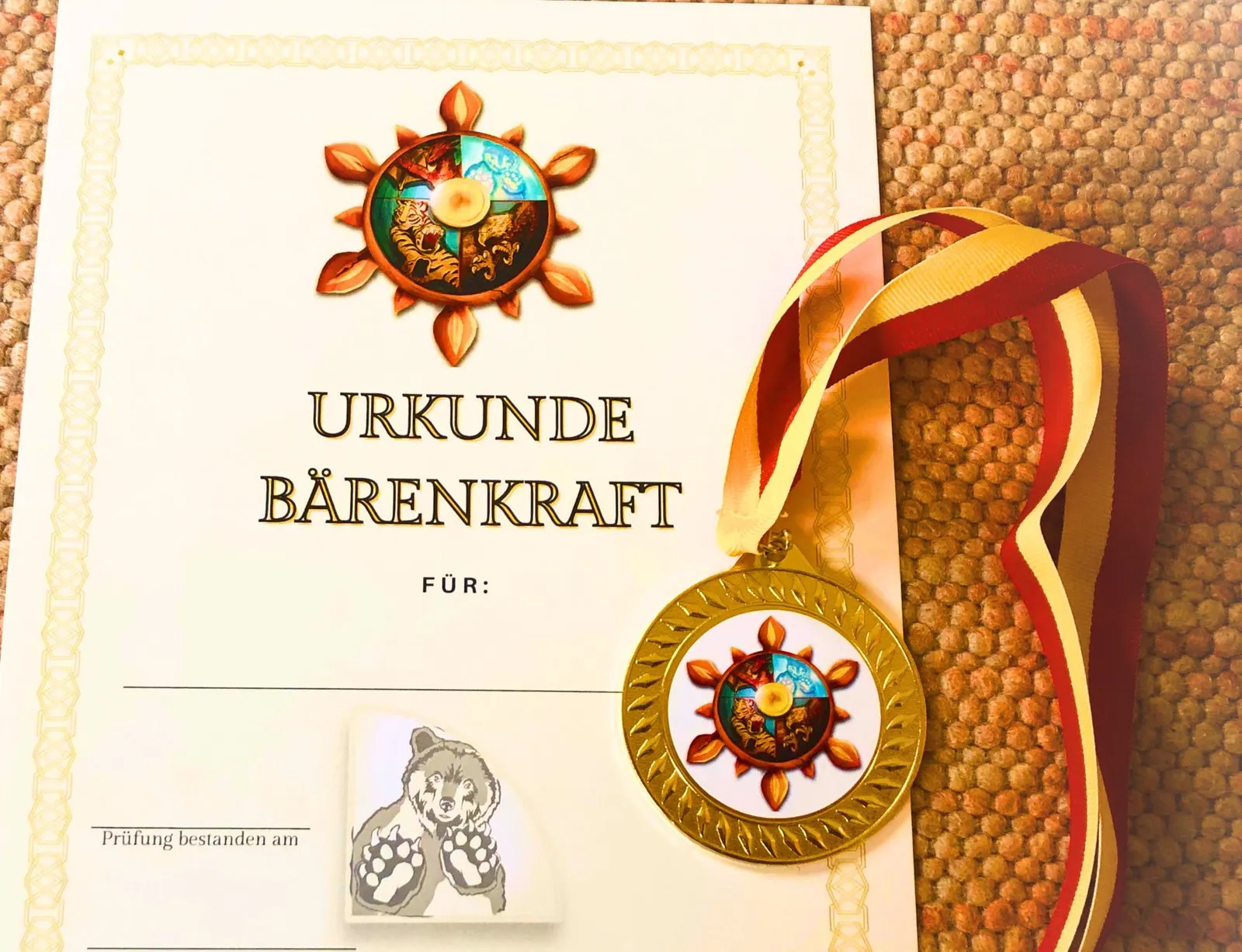Eine Urkunde mit der Aufschrift „Urkunde Bärenkraft“ und dem Rad des Kung-Fu, daneben eine goldene Medaille an rot-goldenem Band.